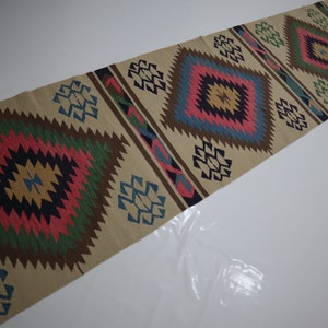 May include: A beige runner rug with a repeating diamond pattern in shades of pink, green, blue, and brown. The rug has a geometric design with a central diamond shape and smaller geometric shapes along the edges. The rug is made of a woven material.