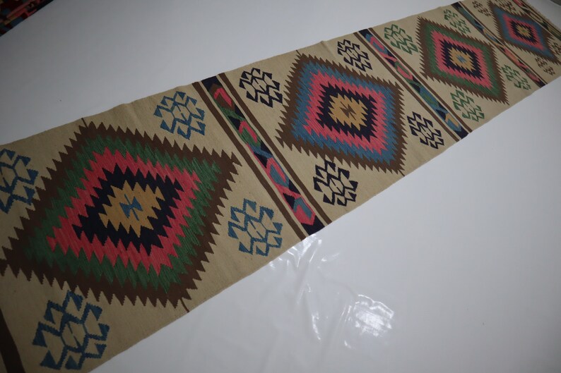 May include: A beige runner rug with a repeating diamond pattern in shades of pink, green, blue, and brown. The rug has a geometric design with a central diamond shape and smaller geometric shapes along the edges. The rug is made of a woven material.