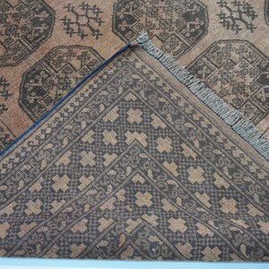May include: Close-up of a brown and black patterned rug. The rug features geometric designs and a detailed border. The corner of the rug is folded back, revealing the underside with a contrasting pattern. The rug has a fringe along one edge.