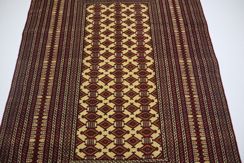 May include: A traditional rug with a cream background and intricate red, black, and yellow geometric patterns. The rug has a rectangular shape and a fringed edge.