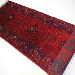 May include: A red runner rug with a traditional Persian design. The rug has a dark blue border with intricate patterns and a central medallion. The rug is rolled up and ready to be unfurled.