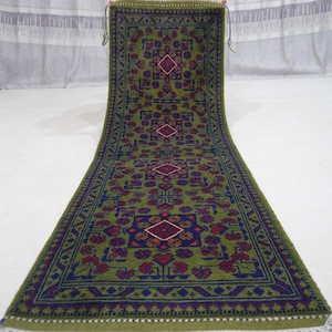 Peut inclure: Un long tapis rectangulaire vert avec un motif géométrique répétitif rouge, bleu et blanc. Le tapis a une bordure avec un motif bleu foncé et des franges blanches aux extrémités. Le tapis est présenté sur une surface blanche.