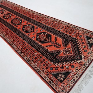 May include: A long, rectangular rug with a central design in shades of orange, black, and brown. The rug features geometric patterns and a fringed edge. The rug's design includes repeating shapes and a central medallion.