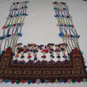 Op de afbeelding: Een kleurrijk textiel met ingewikkelde borduursels en kralenstrengen. Het centrale gedeelte heeft een geometrisch patroon in bruin, rood en zwart, versierd met kwastjes en hangende kralenelementen.