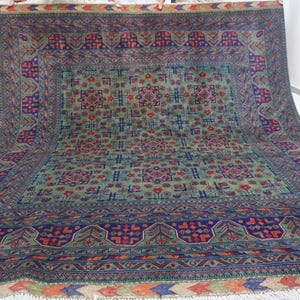 May include: A large, rectangular rug with an intricate, geometric pattern. The rug features a central field of green with red and blue accents. The border is a deep blue with repeating floral and geometric designs. The rug has a fringed edge.