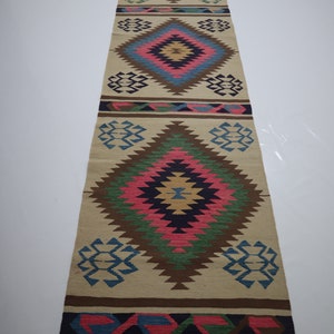 May include: A long, rectangular beige runner rug with a repeating geometric diamond pattern in shades of pink, blue, green, and brown. The rug has a fringed edge and is suitable for a hallway or entryway.