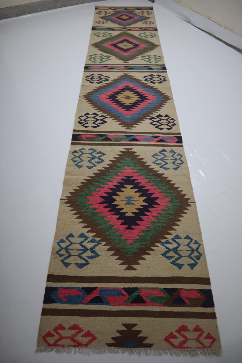 May include: A long, rectangular beige runner rug with a repeating geometric diamond pattern in shades of pink, blue, green, and brown. The rug has a fringed edge and is suitable for a hallway or entryway.