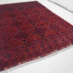 May include: A rectangular, hand-woven rug with a deep red background and intricate geometric patterns in navy blue, green, and cream. The rug features a central diamond design and a detailed border with fringe along the edges.