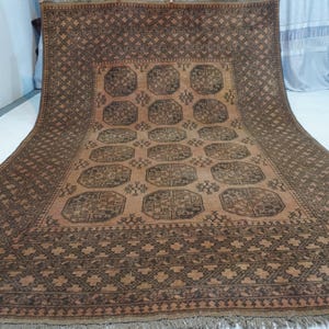 May include: A large, rectangular rug with a detailed geometric pattern in shades of brown and tan. The rug features a central design of repeating medallions and a border with intricate cross-like motifs. The rug has a fringe along the bottom edge.