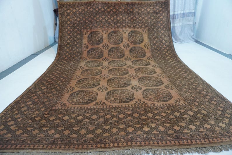 May include: A large, rectangular rug with a detailed geometric pattern in shades of brown and tan. The rug features a central design of repeating medallions and a border with intricate cross-like motifs. The rug has a fringe along the bottom edge.