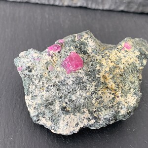 May include: A close-up of a rough, gray rock with several small, pink ruby crystals embedded in it.