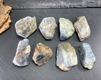 May include: A collection of rough-cut, natural sapphire gemstones. The stones display a mix of blue, gray, and brown hues, with visible textures and patterns. They are arranged on a dark, textured surface.