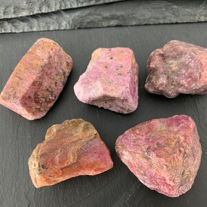 May include: Five rough ruby gemstones in varying shapes and sizes. The stones display a range of pink and reddish-brown hues, with visible crystalline structures. The gemstones are arranged on a dark surface.