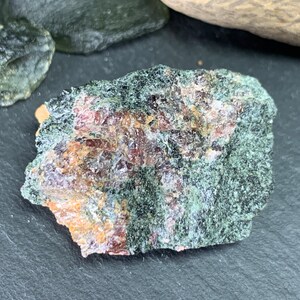 May include: A close-up of a rough, irregularly shaped rock with a dark green exterior and reddish-brown and yellow interior. The rock has a textured surface and appears to be a mineral specimen.