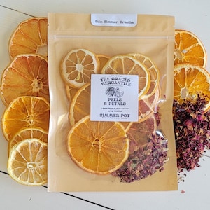 May include: A clear bag filled with dried orange and lemon slices, and rose petals. The bag has a label that reads "PEELS & PETALS" and "Simmer Pot". Sliced oranges and lemons are arranged around the bag.