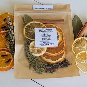 May include: A clear bag filled with dried orange and lemon slices, rosemary, cloves, and cinnamon sticks. The bag has a label that reads "The Graced Mercantile Simply Graced Simmer Pot". The words "Stir. Simmer. Breathe." are printed on the top of the bag.