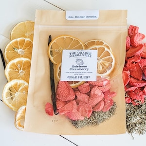 May include: A clear bag filled with dried strawberries, lemon slices, and herbs. The bag has a label that reads "Heirloom Strawberry Simmer Pot" from "The Graced Mercantile." A vanilla bean is also visible. The top of the bag has the text "adr. Simmer. Breathe."