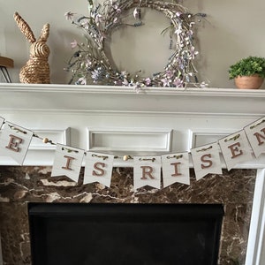 May include: A white wooden banner with the words "He is Risen" in brown letters hangs above a fireplace. The banner is decorated with small green leaves and white beads. A wreath with purple and white flowers is on the mantle above the banner.