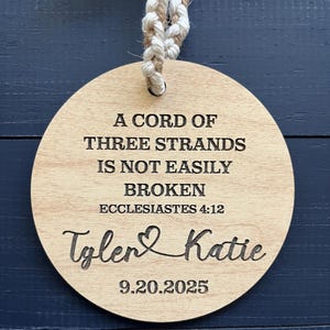 May include: A round wooden ornament with a braided rope hanger. The ornament features engraved text: "A CORD OF THREE STRANDS IS NOT EASILY BROKEN" and "Ecclesiastes 4:12". Below the text are the names "Tyler & Katie" and the date "9.20.2025".