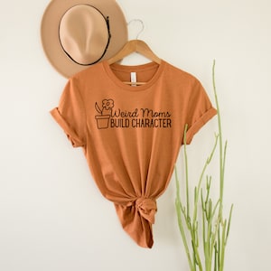 May include: A burnt orange t-shirt with a black graphic of a potted plant and the text "Weird Moms Build Character".