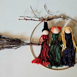 May include: A whimsical Halloween decoration featuring three yarn dolls dressed as witches, each with a unique hair color and dress. They are arranged within a gold hoop, with a broom and decorative branch.