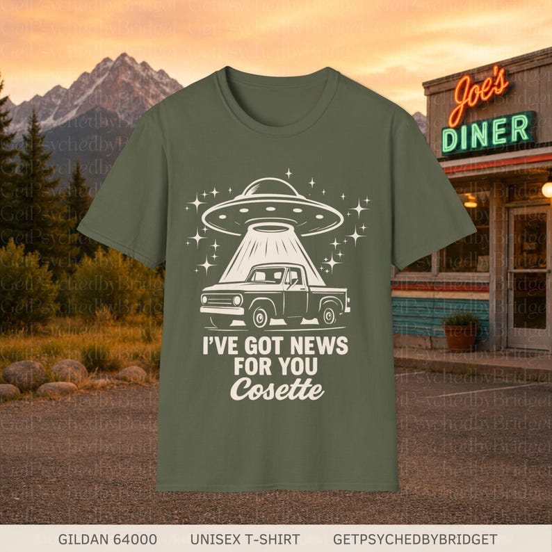 May include: An olive green t-shirt featuring a white graphic of a pickup truck being abducted by a UFO. The design includes the text "I'VE GOT NEWS FOR YOU Cosette" in a bold, stylized font, surrounded by small star shapes on the front of the shirt.