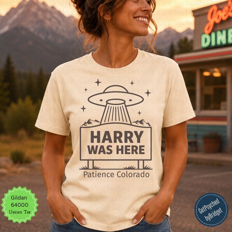 May include: A cream-colored short-sleeve t-shirt featuring a dark brown graphic of a UFO hovering over a rectangular sign that reads HARRY WAS HERE. Below the sign, the text reads Patience Colorado, surrounded by small star icons.