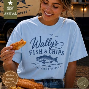 Könnte beinhalten: Hellblaues Unisex-T-Shirt mit dem Text "Wally's Fish & Chips" und einer Fischgrafik. Das Shirt sagt auch "Fritters & Coffee". Das Shirt ist ein Gildan 64000 Unisex Tee.
