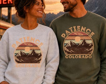 May include: A light gray crewneck sweatshirt and an olive green crewneck sweatshirt, both featuring a graphic print of mountains, a sun, and a UFO with the text PATIENCE COLORADO. The sweatshirts are Gildan 185000 unisex models.