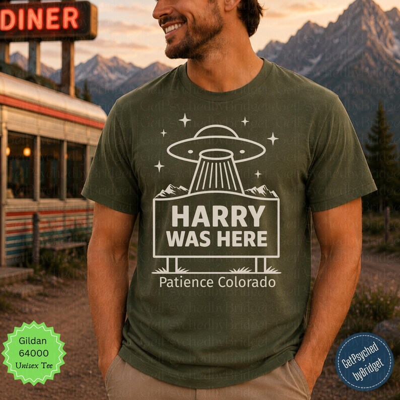 May include: An olive green short-sleeve t-shirt featuring a white graphic of a UFO hovering over a sign that reads HARRY WAS HERE and Patience Colorado. The design includes small star icons and mountain silhouettes above the rectangular sign.