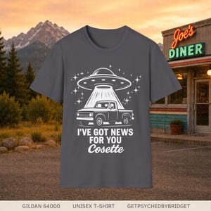 May include: A charcoal gray short-sleeve t-shirt featuring a white graphic of a pickup truck being abducted by a UFO. The text reads I'VE GOT NEWS FOR YOU Cosette. The design includes star accents and a beam of light shining down on the truck.