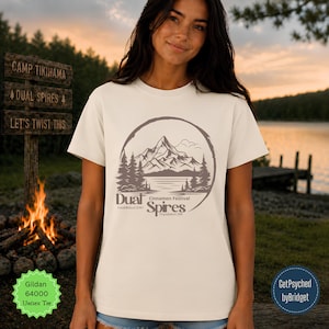 Op de afbeelding: Een crèmekleurig T-shirt met korte mouwen en een donkerbruine cirkelvormige afbeelding van een bergketen, dennenbomen en een meer. De tekst luidt Dual Spires, Cinnamon Festival, Established 2016 en Population 288 in een gestileerd lettertype.