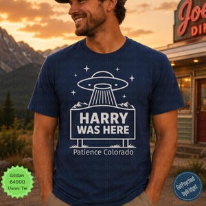 May include: A dark blue short-sleeve t-shirt featuring a white graphic of a UFO hovering over a rectangular sign that reads HARRY WAS HERE with the text Patience Colorado below it, surrounded by small star icons and mountain silhouettes.