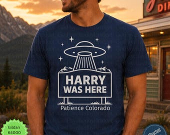 May include: A dark blue short-sleeve t-shirt featuring a white graphic of a UFO hovering over a rectangular sign that reads HARRY WAS HERE with the text Patience Colorado below it, surrounded by small star icons and mountain silhouettes.