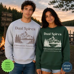May include: A light gray crewneck sweatshirt and a dark green crewneck sweatshirt, both featuring a graphic of mountains, trees, and a lake with the text Dual Spires Cinnamon Festival Established 2010 Population 288 printed on the front.