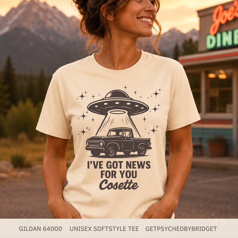 May include: A cream-colored short-sleeve t-shirt featuring a dark graphic of a pickup truck being abducted by a UFO. The design includes star accents and the text I'VE GOT NEWS FOR YOU Cosette printed in a bold, dark font on the front of the shirt.
