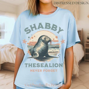 May include: Pale blue t-shirt with a distressed seal design. The graphic includes the words "SHABBY THESEALION NEVER FORGET" in a vintage style. The shirt has short sleeves and a crew neck, suitable for casual wear.