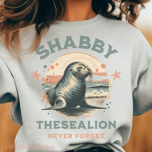 Può includere: Una felpa grigia chiaro con una grafica in stile vintage di un leone marino su una spiaggia. La grafica include il testo "Shabby The Sealion Never Forget" e "Get Psyched by Bridget".