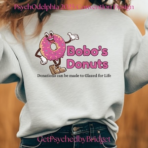 May include: A white sweatshirt featuring a pink and brown cartoon donut character holding a sign that reads "Bobo's Donuts". The text "Donations can be made to Glazed for Life" is below the character. The text "GetPsychedbyBridget" is at the bottom of the sweatshirt.