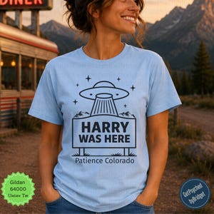 May include: A light blue short-sleeve t-shirt featuring a black graphic of a UFO hovering over a rectangular sign that reads HARRY WAS HERE with the text Patience Colorado below it, surrounded by small star icons and mountain silhouettes.
