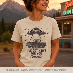 May include: A beige short-sleeve t-shirt featuring a dark brown graphic of a pickup truck being abducted by a UFO. The design includes star accents and the text "I'VE GOT NEWS FOR YOU Cosette" printed in a bold, dark brown sans-serif font.