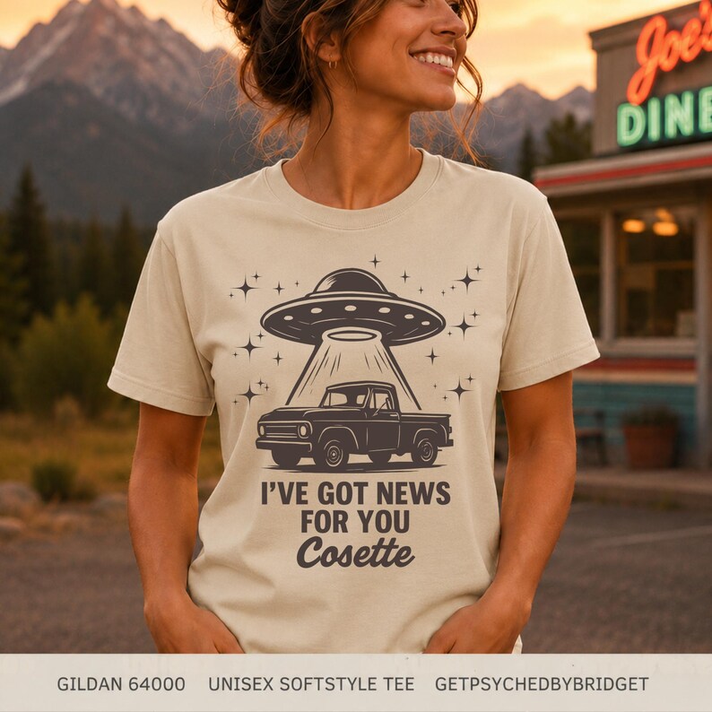 May include: A beige short-sleeve t-shirt featuring a dark brown graphic of a pickup truck being abducted by a UFO. The design includes star accents and the text "I'VE GOT NEWS FOR YOU Cosette" printed in a bold, dark brown sans-serif font.