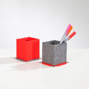 Könnte beinhalten: Zwei Filz-Bleistifthalter, einer rot und einer grau. Der graue Halter enthält drei Stifte, einen rosa, einen orangefarbenen und einen silbernen.
