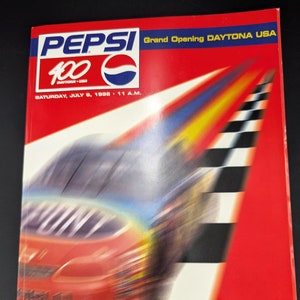 May include: A red and blue Pepsi logo with the text "Pepsi 400 Daytona USA" and "Grand Opening Daytona USA". The text "Saturday, July 6, 1998, 11 A.M." is below the logo. The image also features a red race car speeding down a track with a checkered finish line. The text "Daytona International Speedway" and "Special Commemorative Edition" is at the bottom of the image.