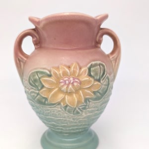 May include: A small, decorative vase with a floral design. The vase features a two-tone color scheme, with a pink upper section and a light blue base. A yellow flower with pink accents is the focal point.
