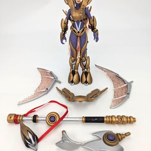 May include: A detailed action figure of a female warrior in gold and purple armor, with multiple accessories including wings, a staff, and weapons. The figure has a helmet and long, flowing hair. The accessories are silver and gold.