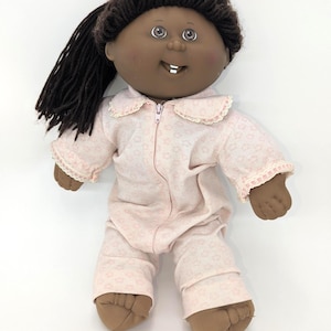 May include: A vintage doll with dark skin, brown hair, and a smiling expression. The doll is wearing a light pink onesie with a floral pattern and a zipper closure. The onesie has lace trim around the collar and cuffs.
