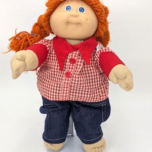 May include: A vintage Cabbage Patch Kids doll with orange yarn hair styled in braids. The doll wears a red and white checkered shirt, red sleeves, and dark blue denim pants. The doll has blue eyes and a smiling expression.