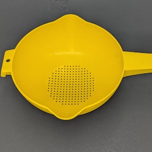 May include: A bright yellow plastic colander featuring a round bowl with a perforated base for draining. It includes a long handle with a small circular hole at the end and a smaller tab on the opposite side, also with a circular hole for hanging.