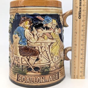 May include: A ceramic beer stein with a detailed relief scene of people at a table, with the words "1/2 GALLON OF ALE" at the base. The stein has a brown handle and a brown rim. The stein is approximately 7 inches tall.